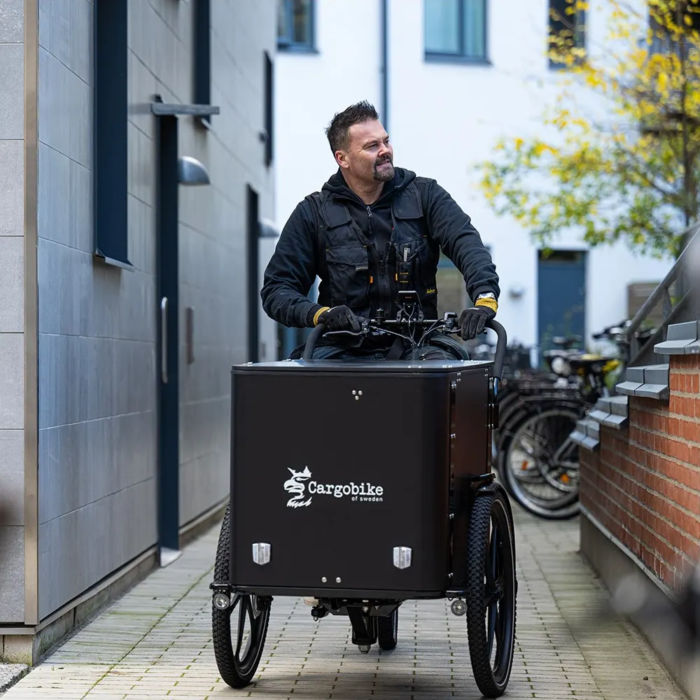 Man cyklar en svart Cargobike Delight Box mellan bostadshus på en stenlagd gång